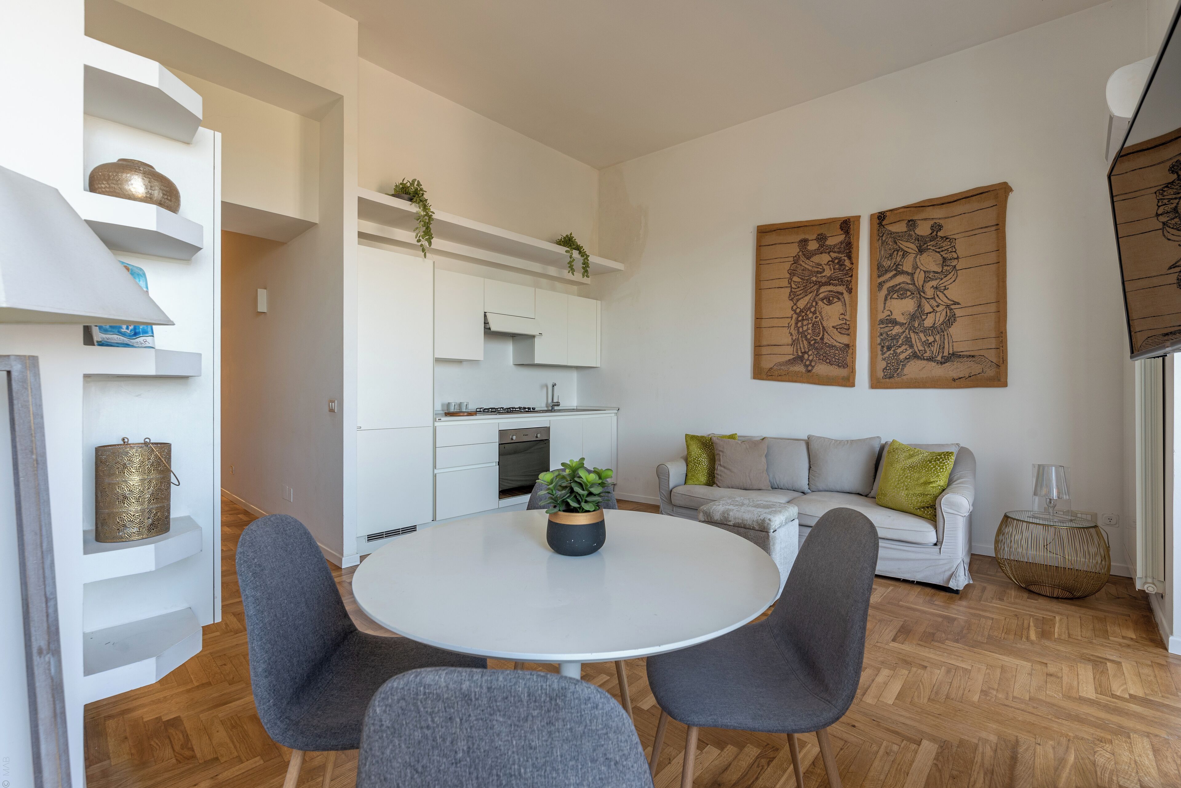 A modern living room with a round white table, grey chairs, a grey sofa with green and beige cushions, and a white kitchen in the background.