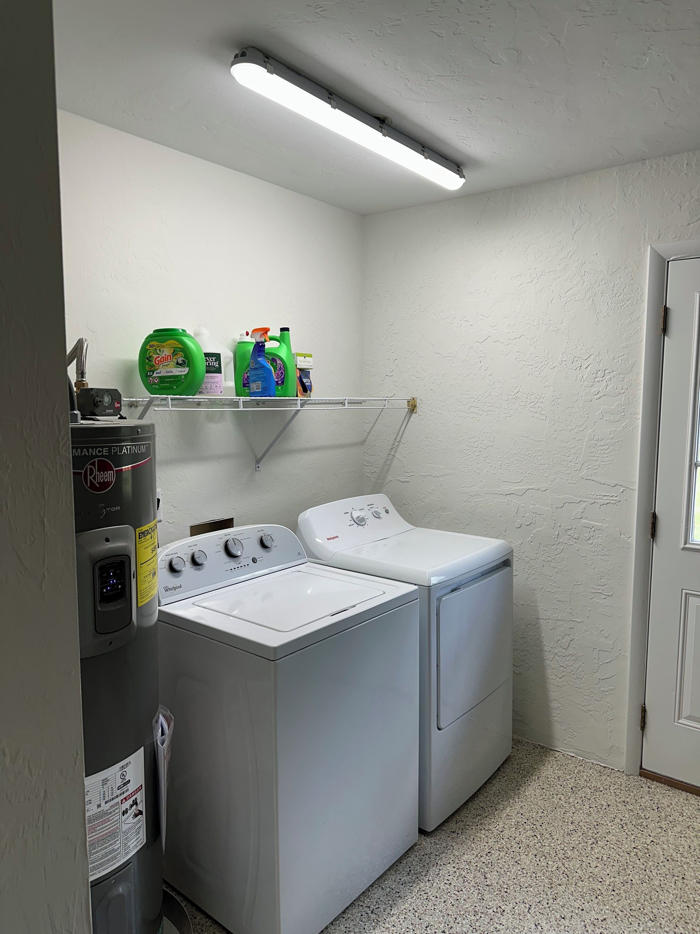A laundry room with two front-loading washers and dryers, a shelf with cleaning supplies, and a water heater.