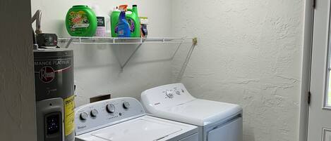 A laundry room with two front-loading washers and dryers, a shelf with cleaning supplies, and a water heater.