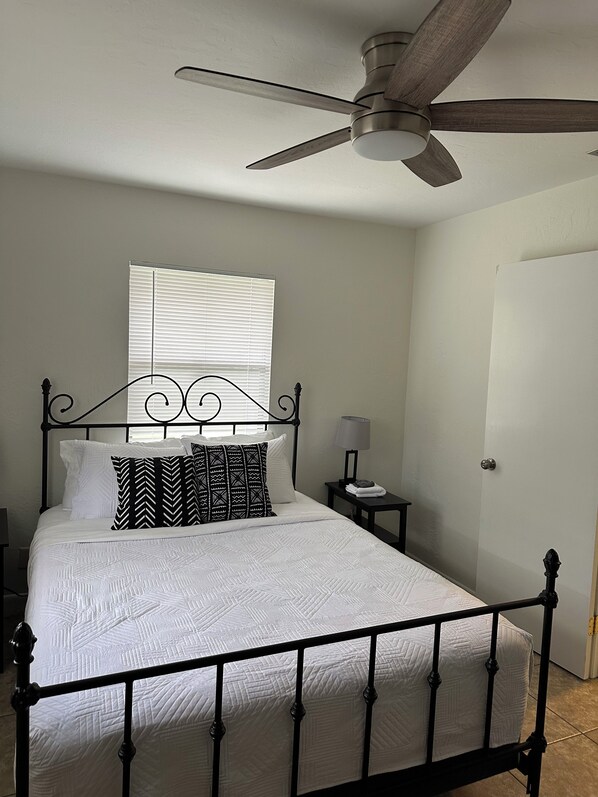 A neatly made bed with a black metal frame, a ceiling fan, a bedside table with a lamp, and a window with blinds.