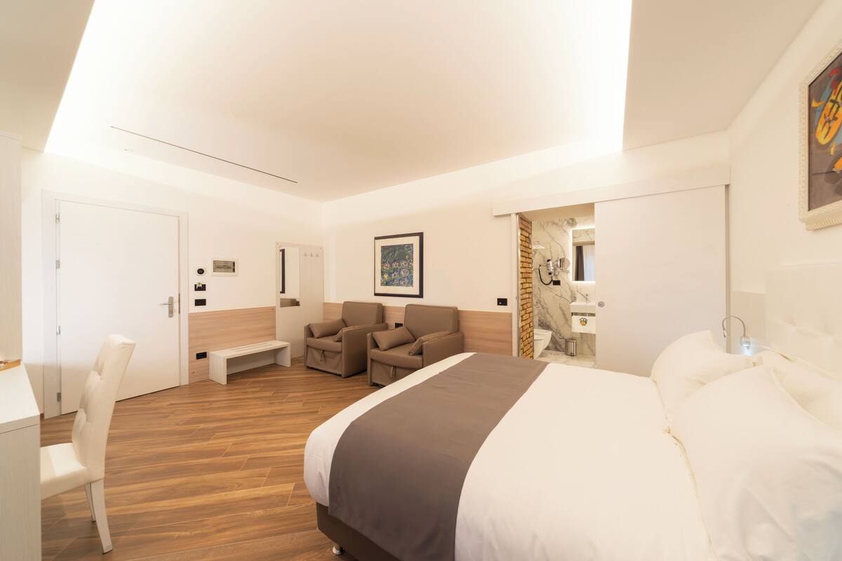 A modern hotel room with a large bed, a seating area with two sofas, a desk, and a bathroom visible through an open door.