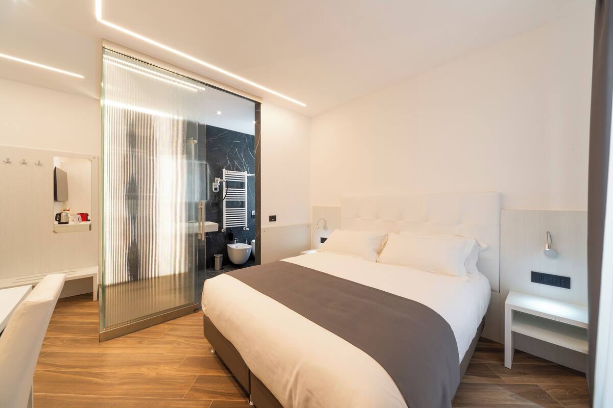 A modern hotel room with a large bed, a glass partition wall, a small bathroom, and a bedside table.