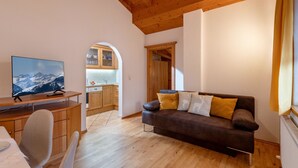 A living room with a wooden ceiling, a sofa, a TV, and a kitchen in the background. - Apartment 'Lisa' with Mountain View, Balcony and Wi-Fi (Fügen)