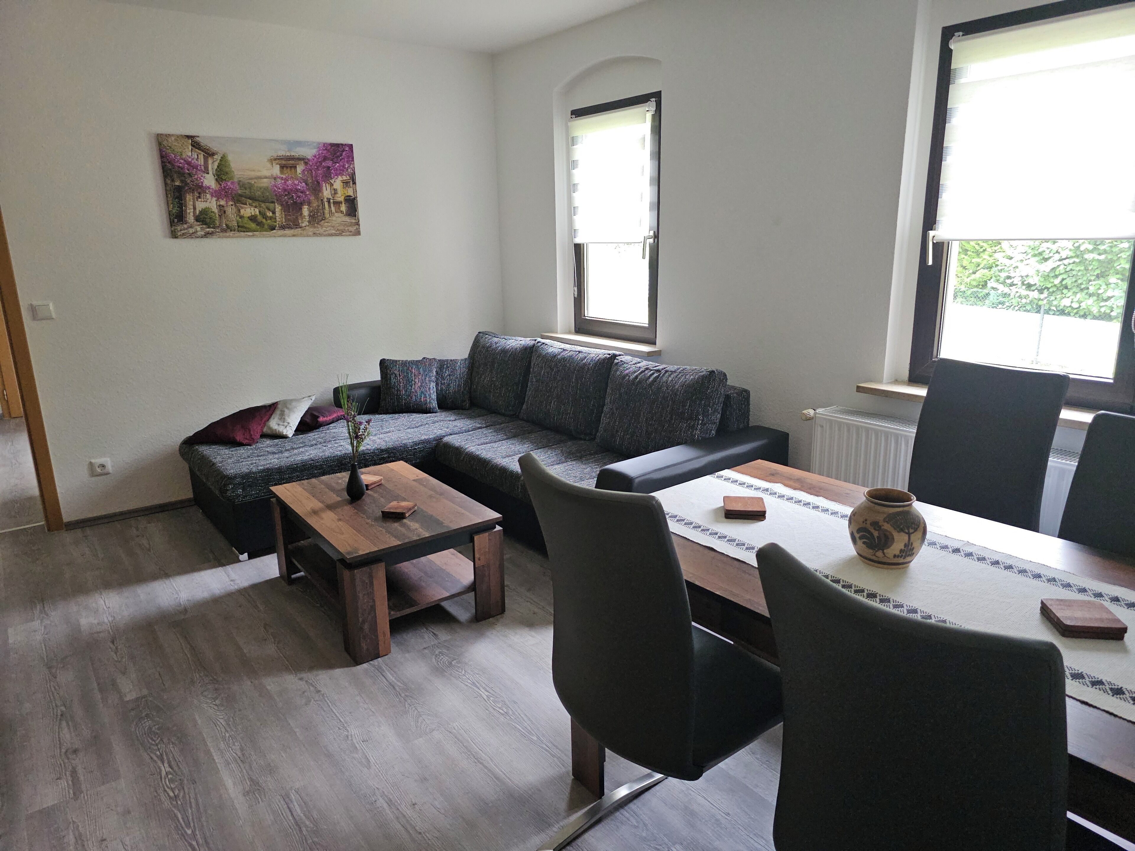 Ein Wohnzimmer mit einem Ecksofa, einem Couchtisch und einem Esstisch mit Stühlen. An der Wand hängt ein Gemälde und das Fenster bietet Blick auf Grünflächen.