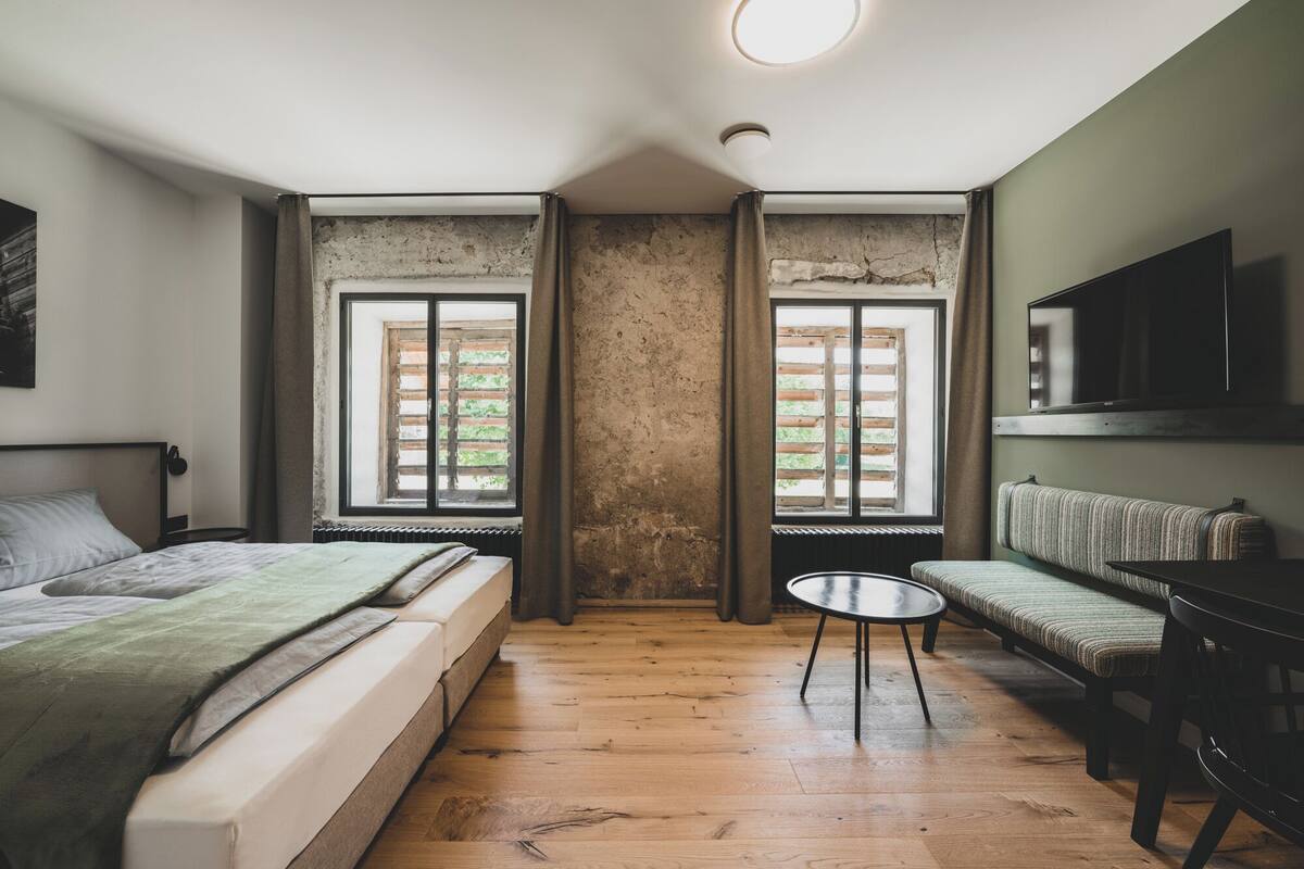 A room with a bed, a sofa, a TV, and two windows with shutters.