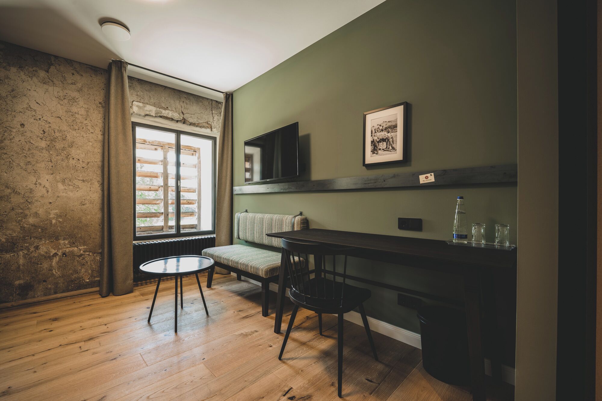 Una habitación con piso de madera, una ventana con cortinas, un banco, una mesa y un cuadro enmarcado en la pared.