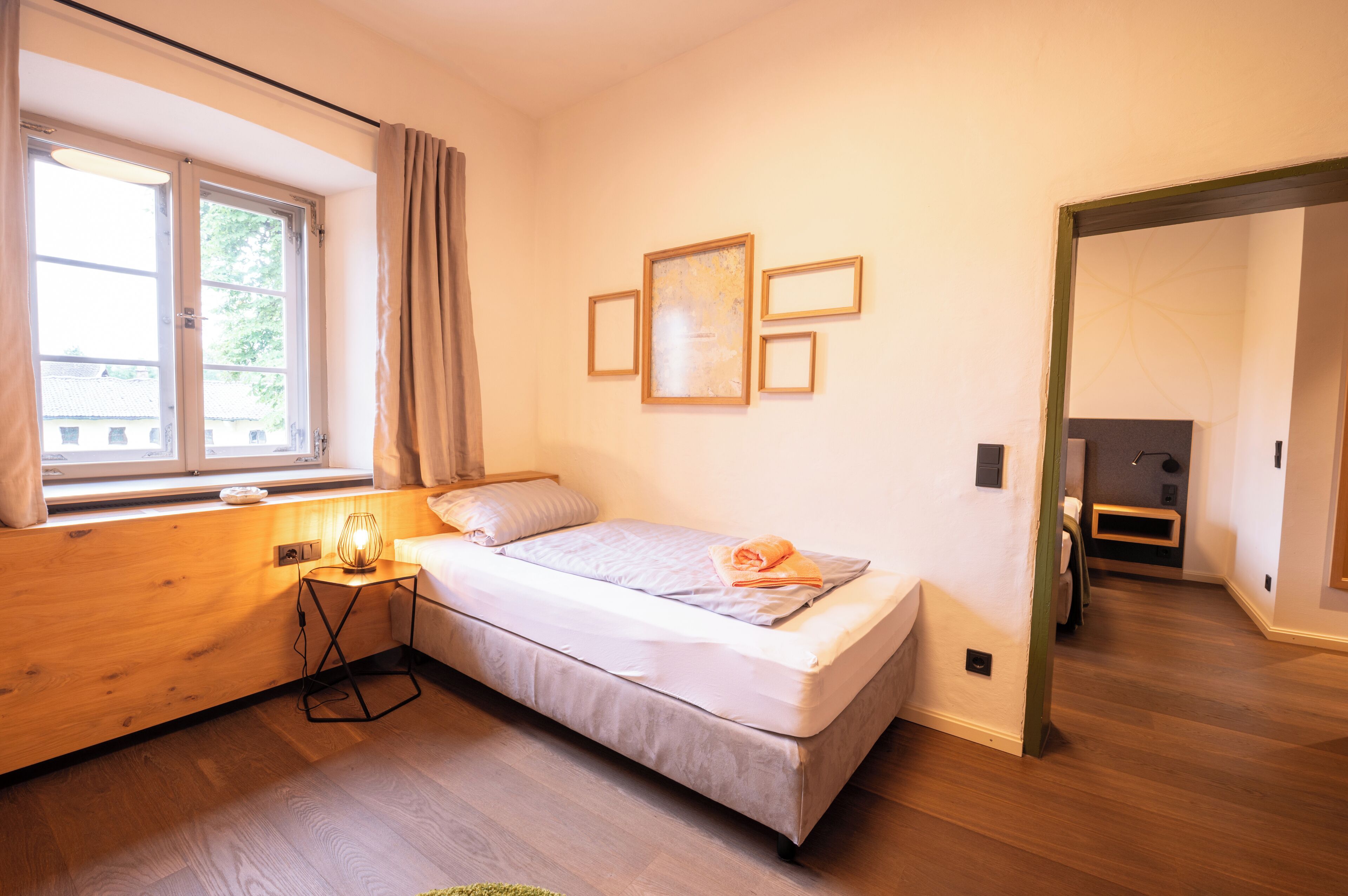 Un dormitorio con cama, mesita de noche con lámpara, ventana con cortinas y un cuadro en la pared.