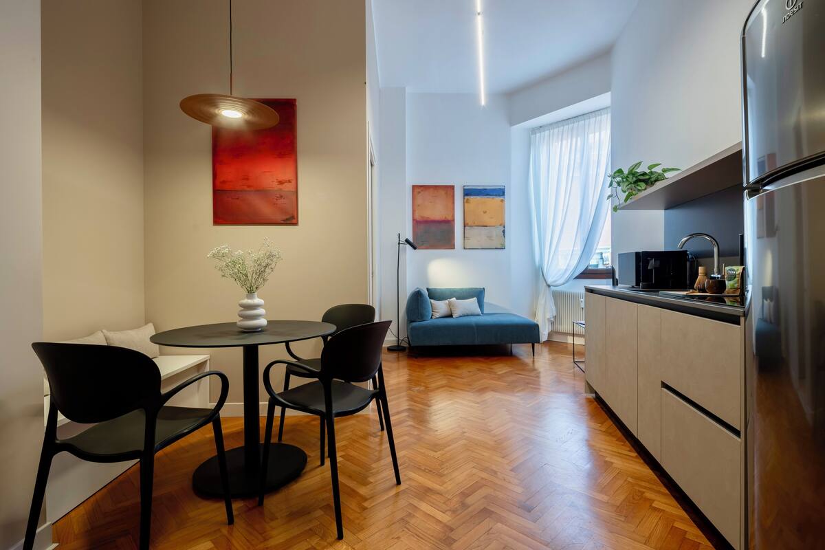 A modern kitchen with a refrigerator, a dining area with a round table and chairs, and a living area with a sofa and artwork on the wall.
