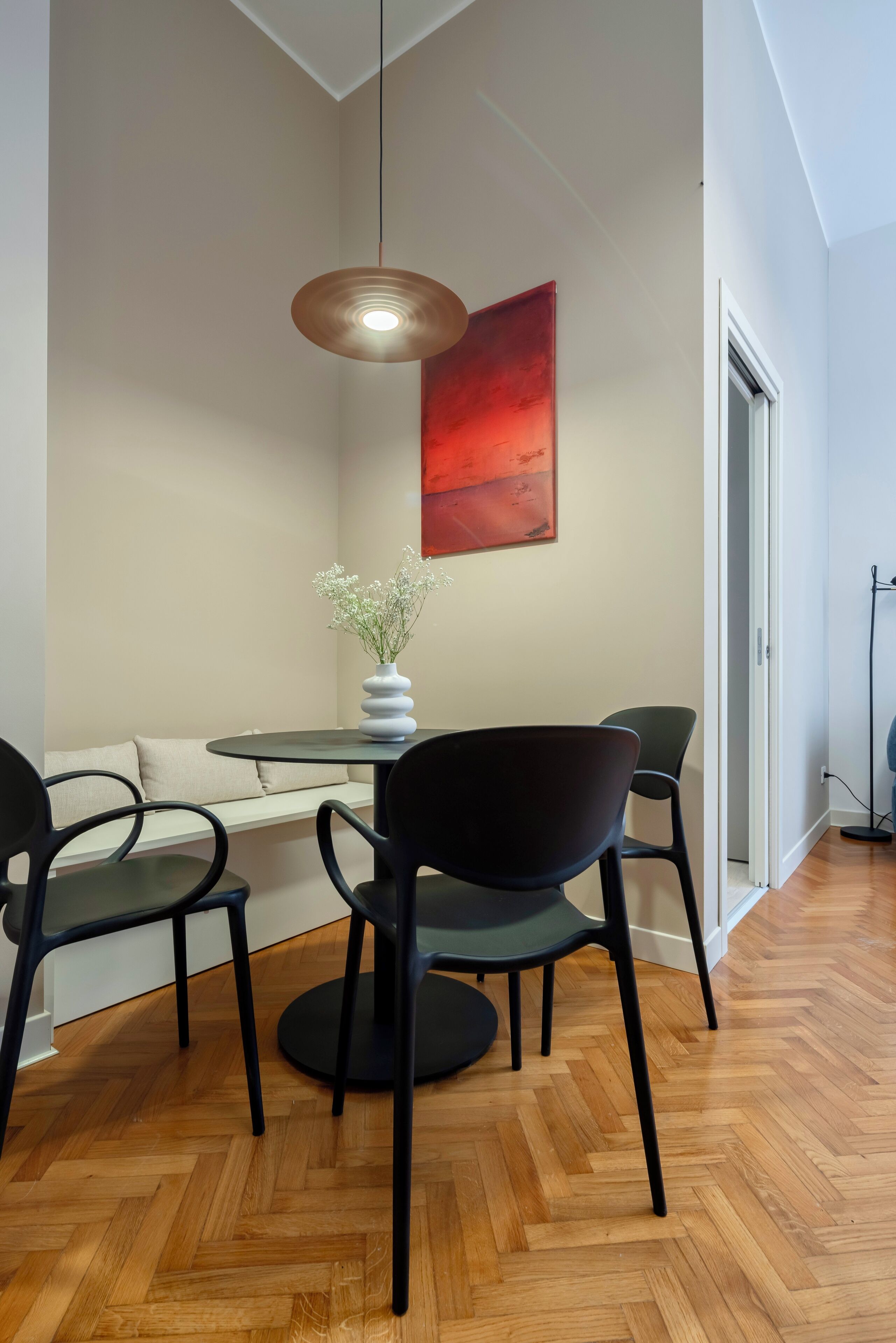 Un coin repas comprenant une table ronde, deux chaises, un vase avec des fleurs, un tableau abstrait rouge et un luminaire suspendu.