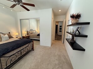 A bedroom with a bed, bedside tables, a ceiling fan, a mirror, and a hallway leading to a bathroom.