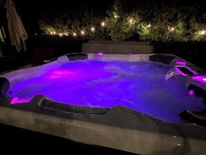 A hot tub with a purple light, surrounded by a hedge and string lights.