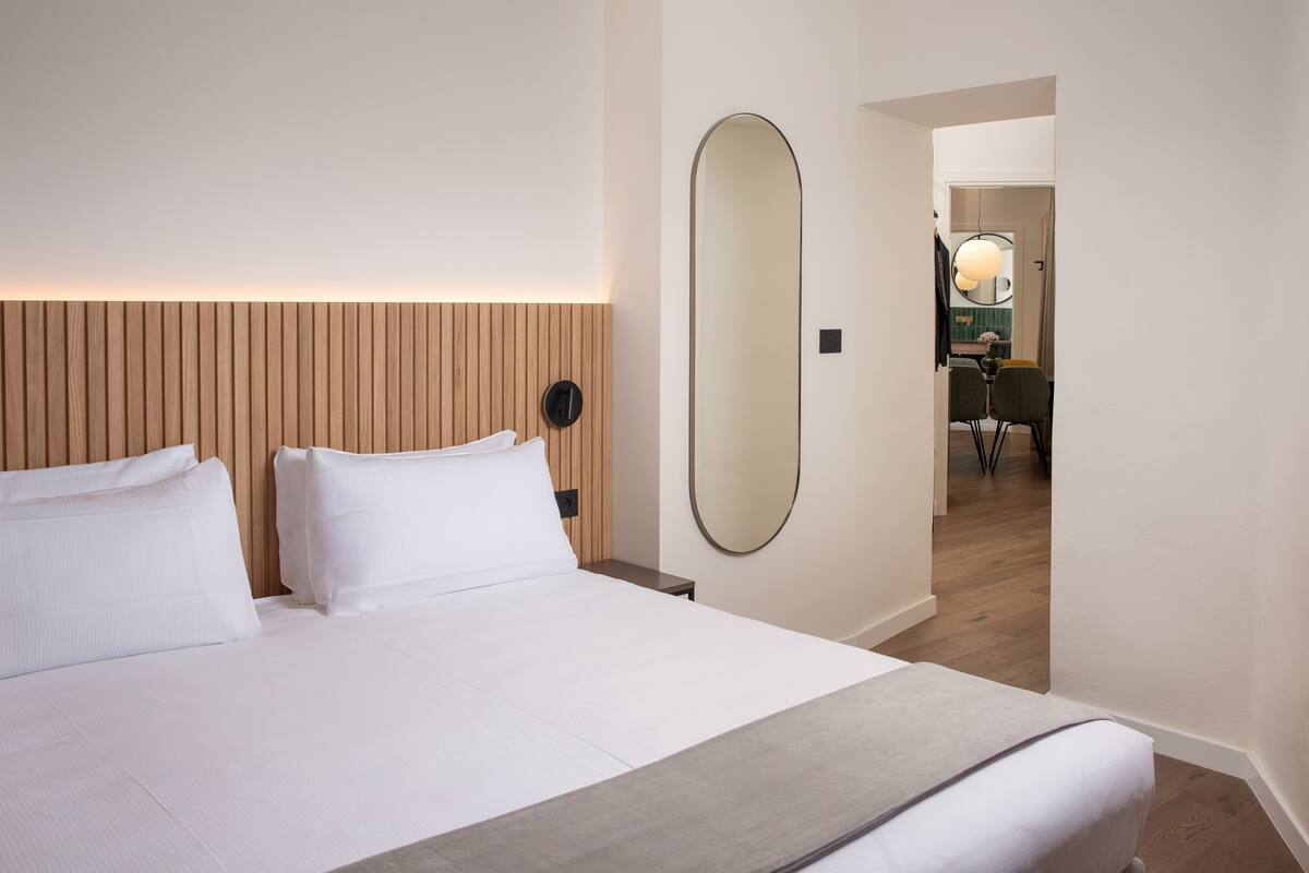A hotel room with a large bed, a wooden headboard, a round mirror, and a view into the dining area.