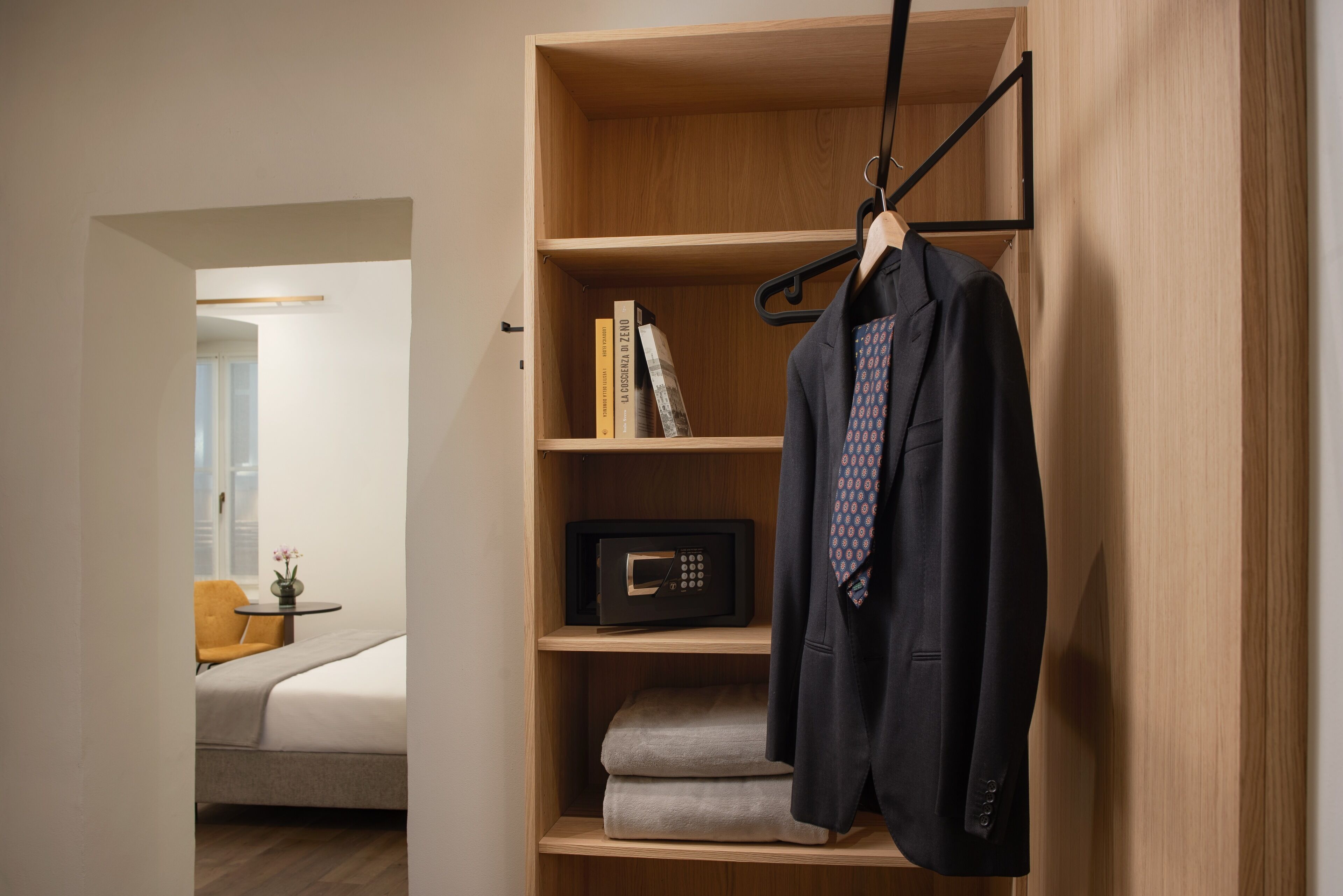A room with a bed, a small table with a vase, and a wooden bookshelf with a coat, a book, and a safe.