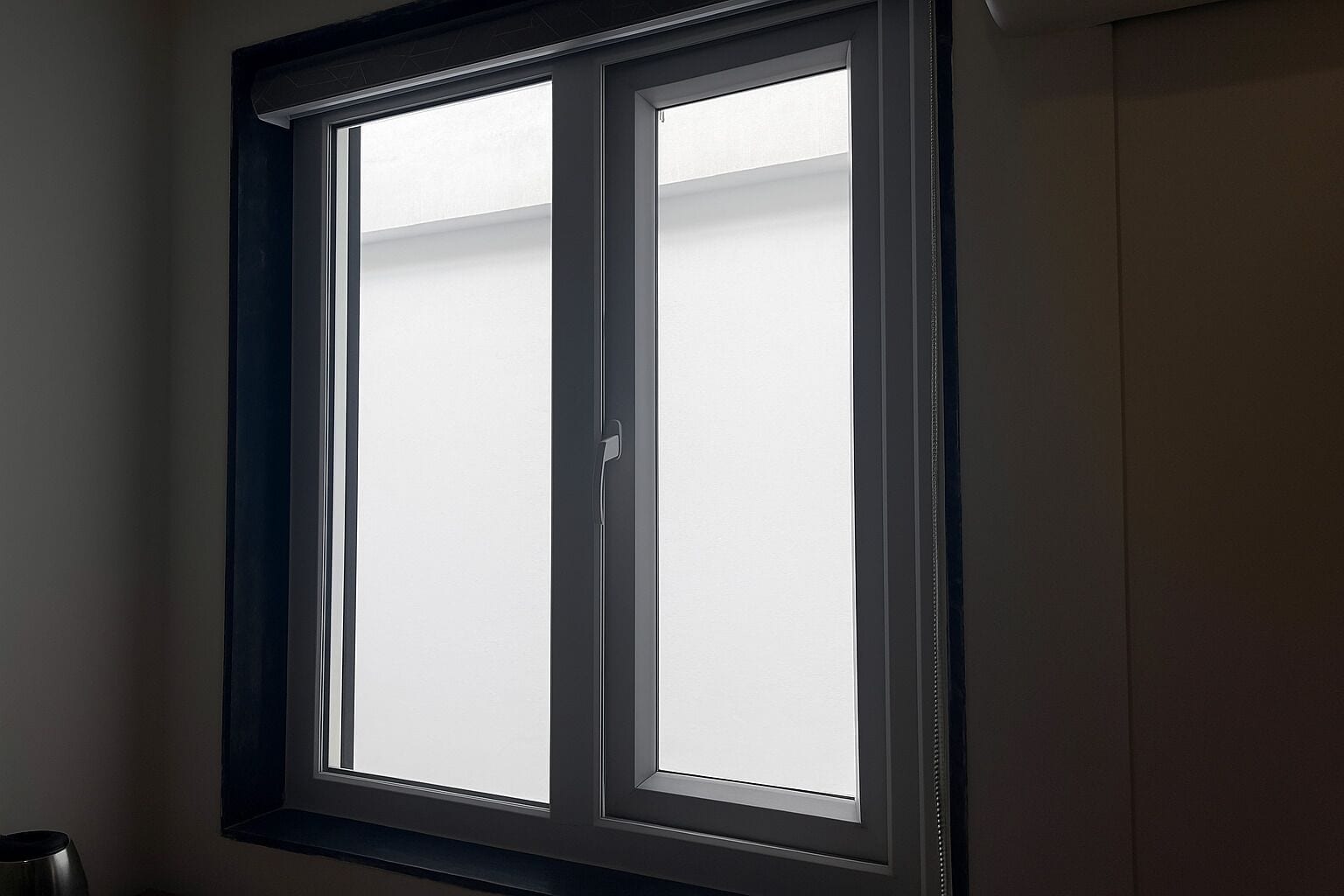 A window with a dark frame and a view of a white wall.