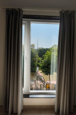 A room with a large window, grey curtains, and a view of a park with trees and a fountain. - Hotel Veronica (Indore)