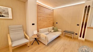 A modern living room with a wooden panel wall, a sofa, a chair, a small table with a lamp, and skis leaning against the wall. - "Madesimo Ski Suite": apartment just steps from the slopes! (Madesimo)