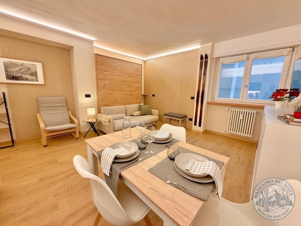 Un salon moderne comprenant un coin repas, un canapé, un fauteuil, une table dressée pour deux personnes et une fenêtre donnant sur un beau paysage.