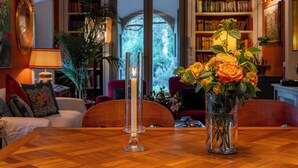 A dining table with a candle and a vase of flowers, surrounded by bookshelves and a view of greenery.