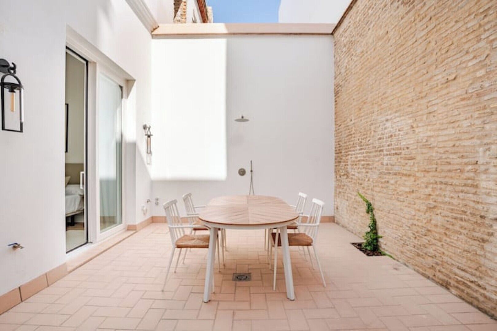 Een terras met een ronde tafel en stoelen, een bakstenen muur en een schuifdeur die leidt naar een kamer.
