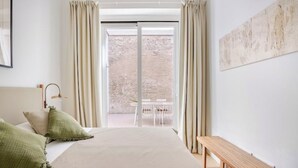 Une chambre avec un grand lit, un banc, une lampe de bureau et une vue sur un mur de briques et un espace extérieur aménagé, visible à travers une porte coulissante.