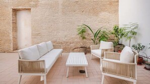 Une terrasse avec un canapé blanc, deux fauteuils en osier, une table blanche et des plantes en pot, le tout adossé à un mur de briques.