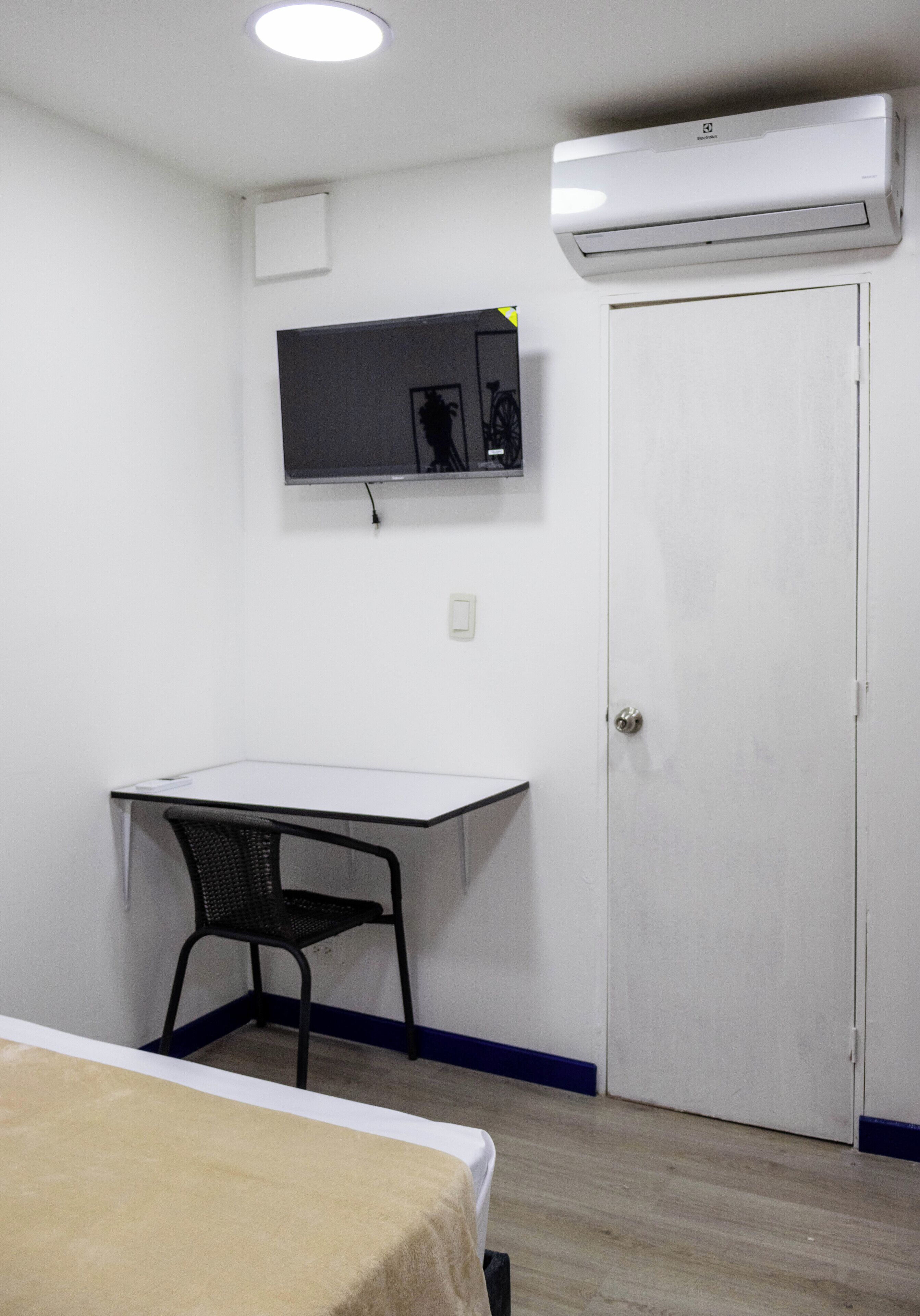 A room with a bed, a desk, a chair, a TV mounted on the wall, and an air conditioning unit.