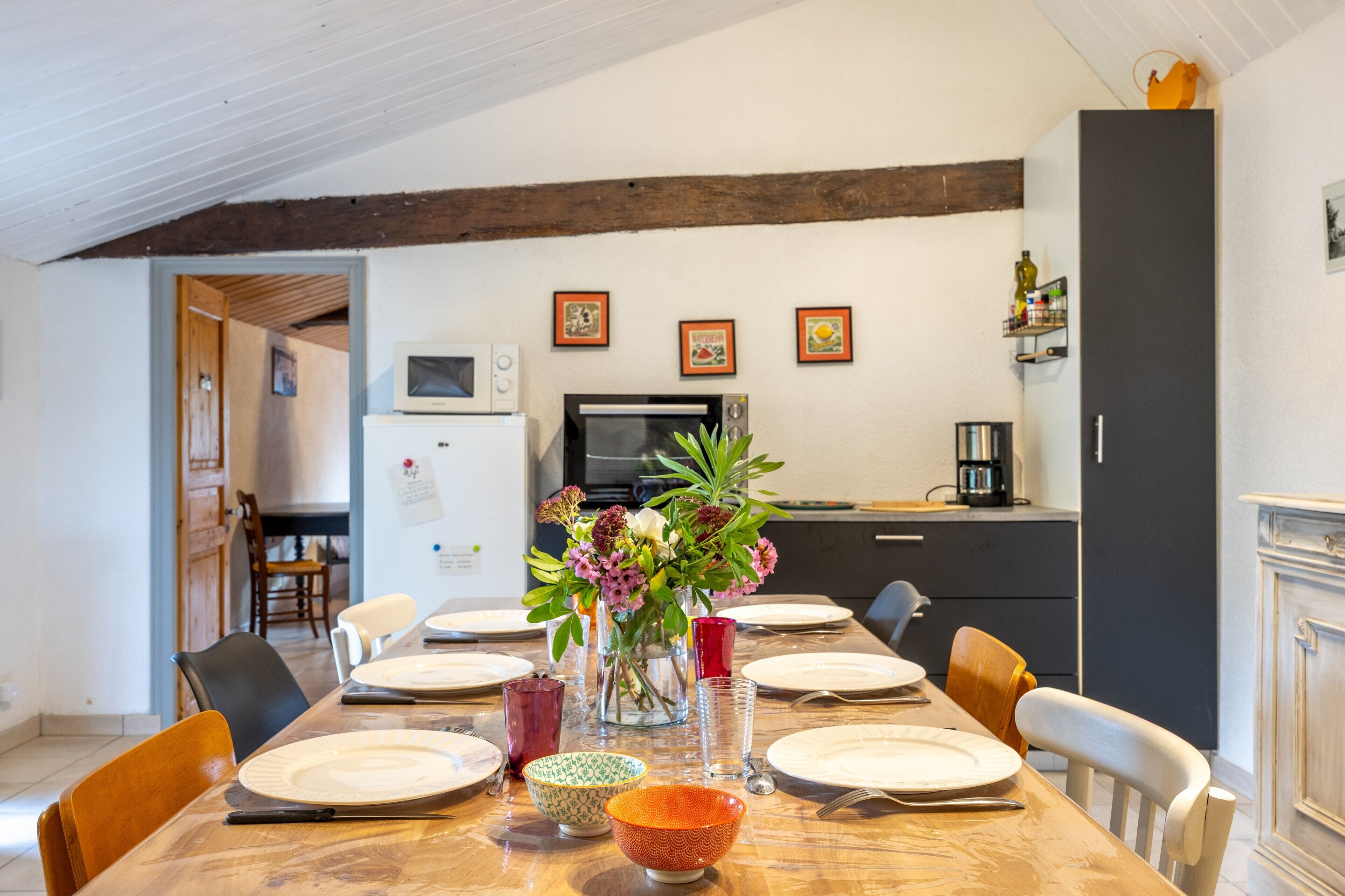 A dining area with a wooden table set for four, surrounded by chairs, with a kitchen in the background.
