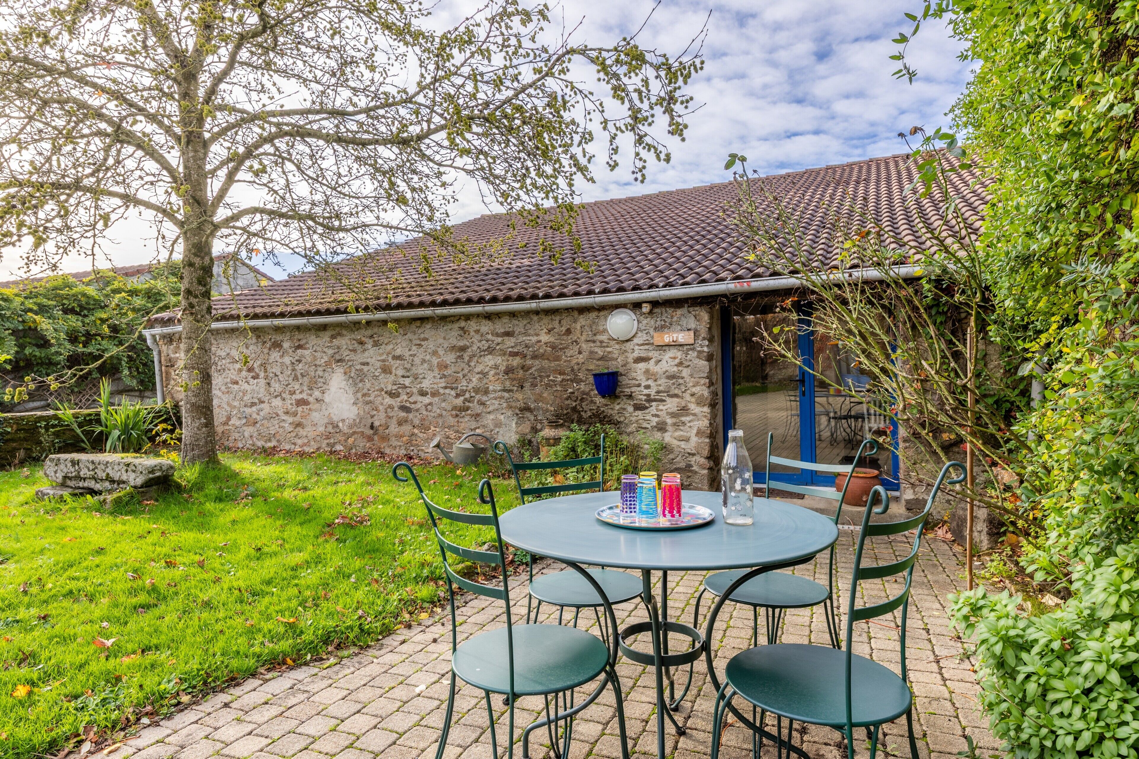 A stone building with a tiled roof, a round table with chairs, and a blue door.