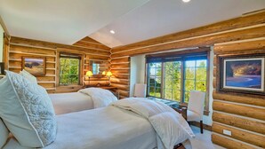 Un dormitorio tipo cabaña de troncos con dos camas, un escritorio y ventanas amplias que ofrecen vistas del exterior.
