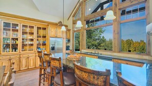 Cocina amplia con armarios de madera, una isla grande con taburetes de bar y vistas a árboles a través de ventanales.