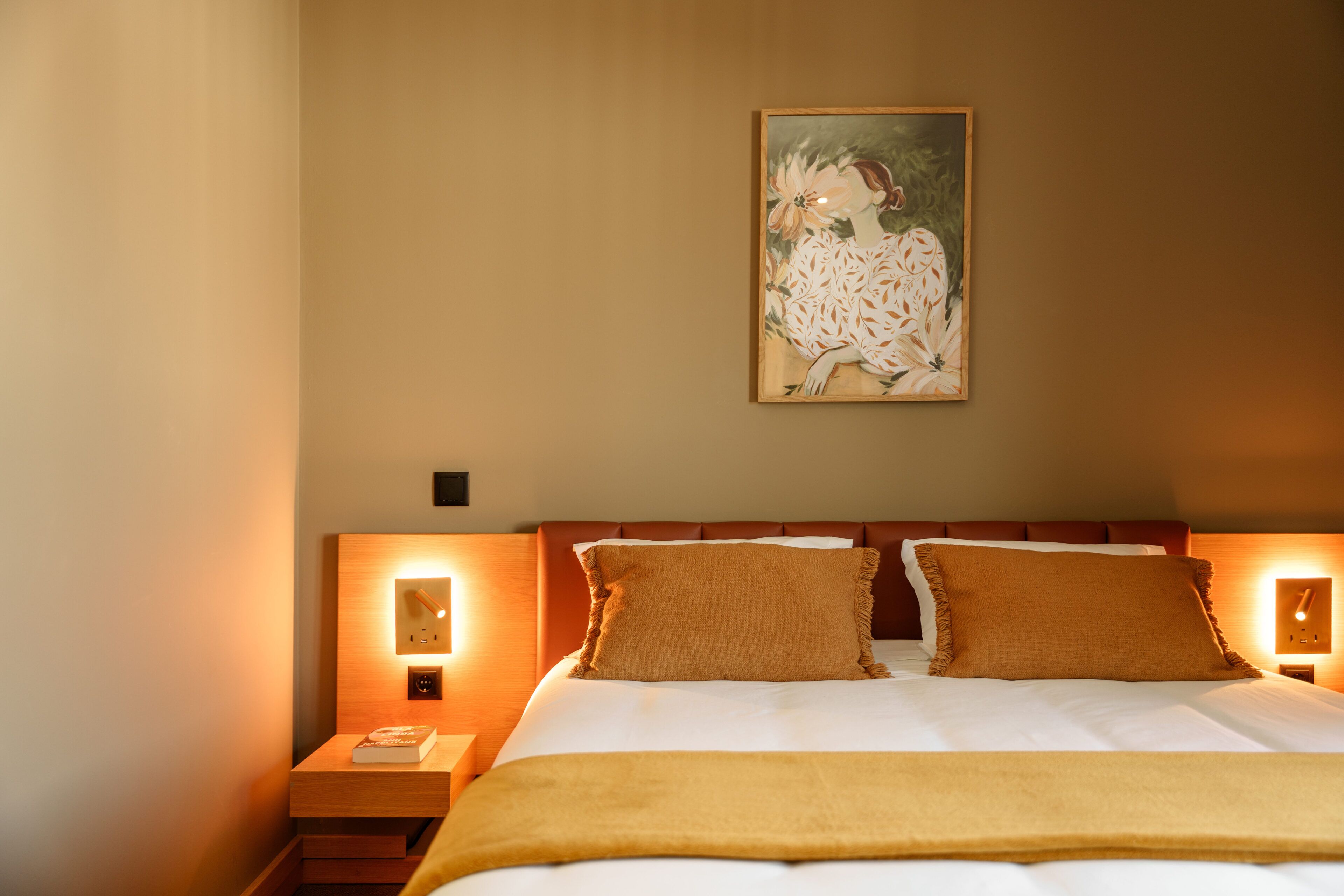 Una cama bien hecha con cabecera, dos lámparas de noche y una obra de arte enmarcada sobre la cama.