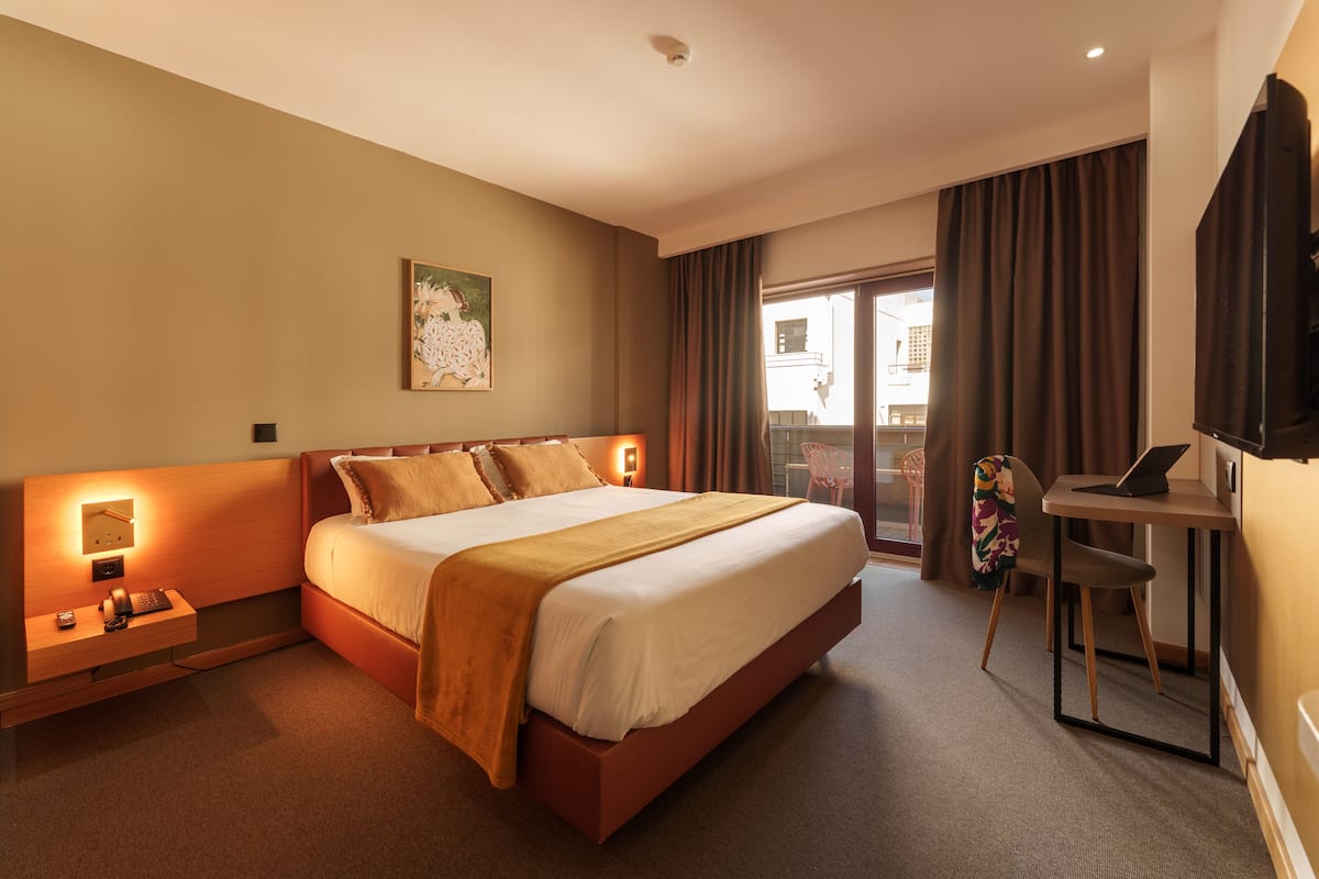 A hotel room with a large bed, bedside table with a lamp, a desk with a laptop, and a window with curtains.