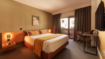 Quarto de hotel com uma cama grande, mesa de cabeceira com abajur, escrivaninha com um laptop e janela com cortinas.