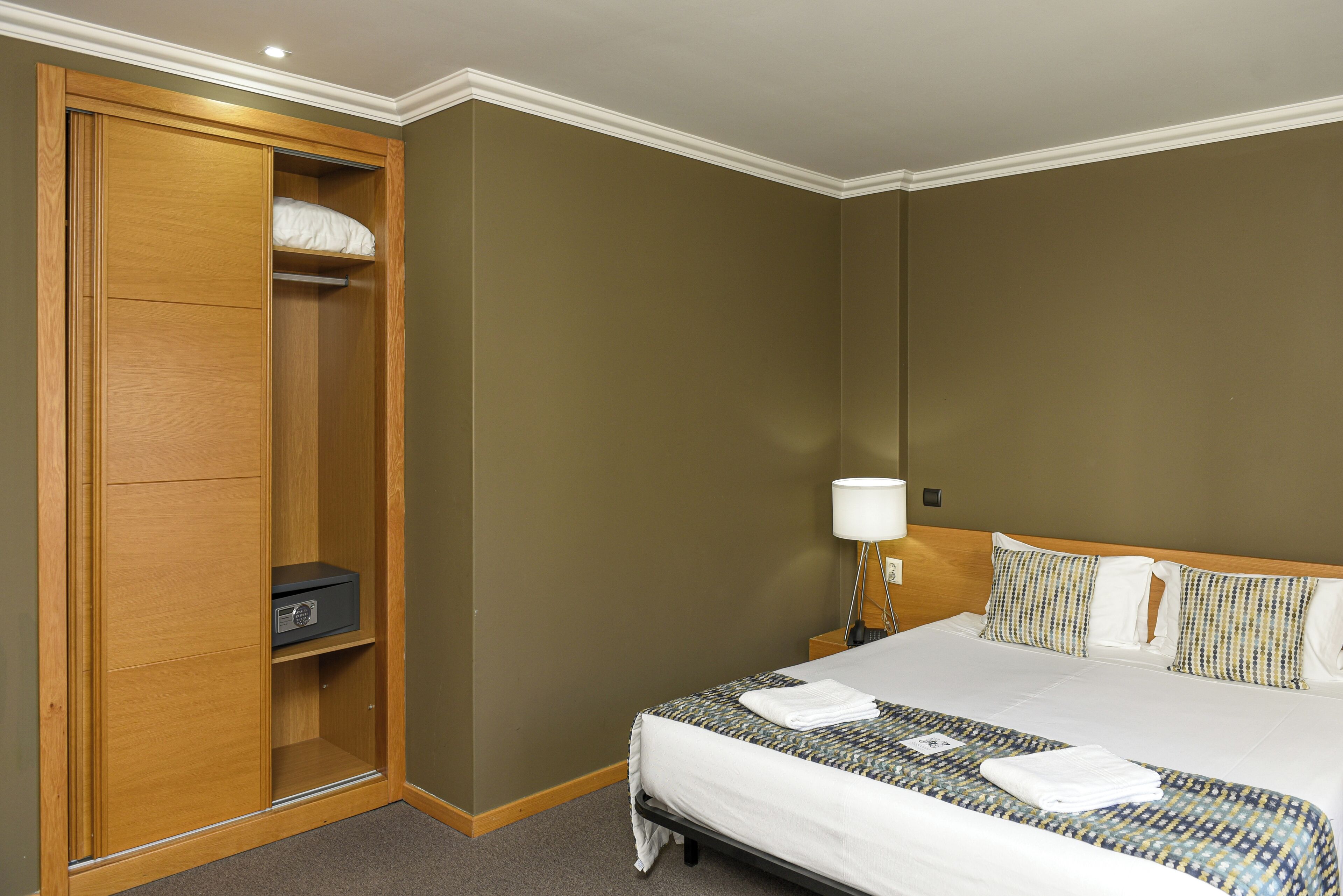 A hotel room with a large bed, a wardrobe with a radio, a bedside table with a lamp, and patterned pillows.
