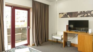 A hotel room with a bed, a desk with a TV, a chair, a window with curtains, and a door leading to a balcony.