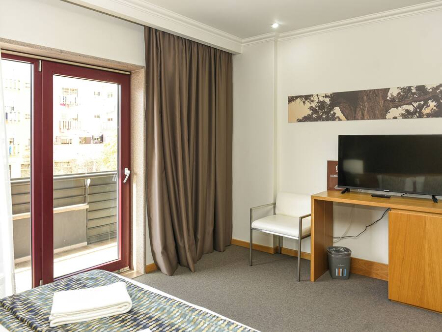 A hotel room with a bed, a desk with a TV, a chair, a window with curtains, and a door leading to a balcony.