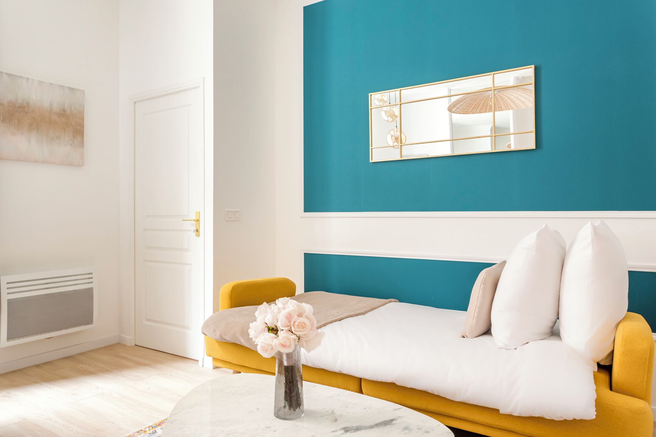 A room with a yellow sofa, white bedding, a vase with flowers, a teal accent wall, and a white door.