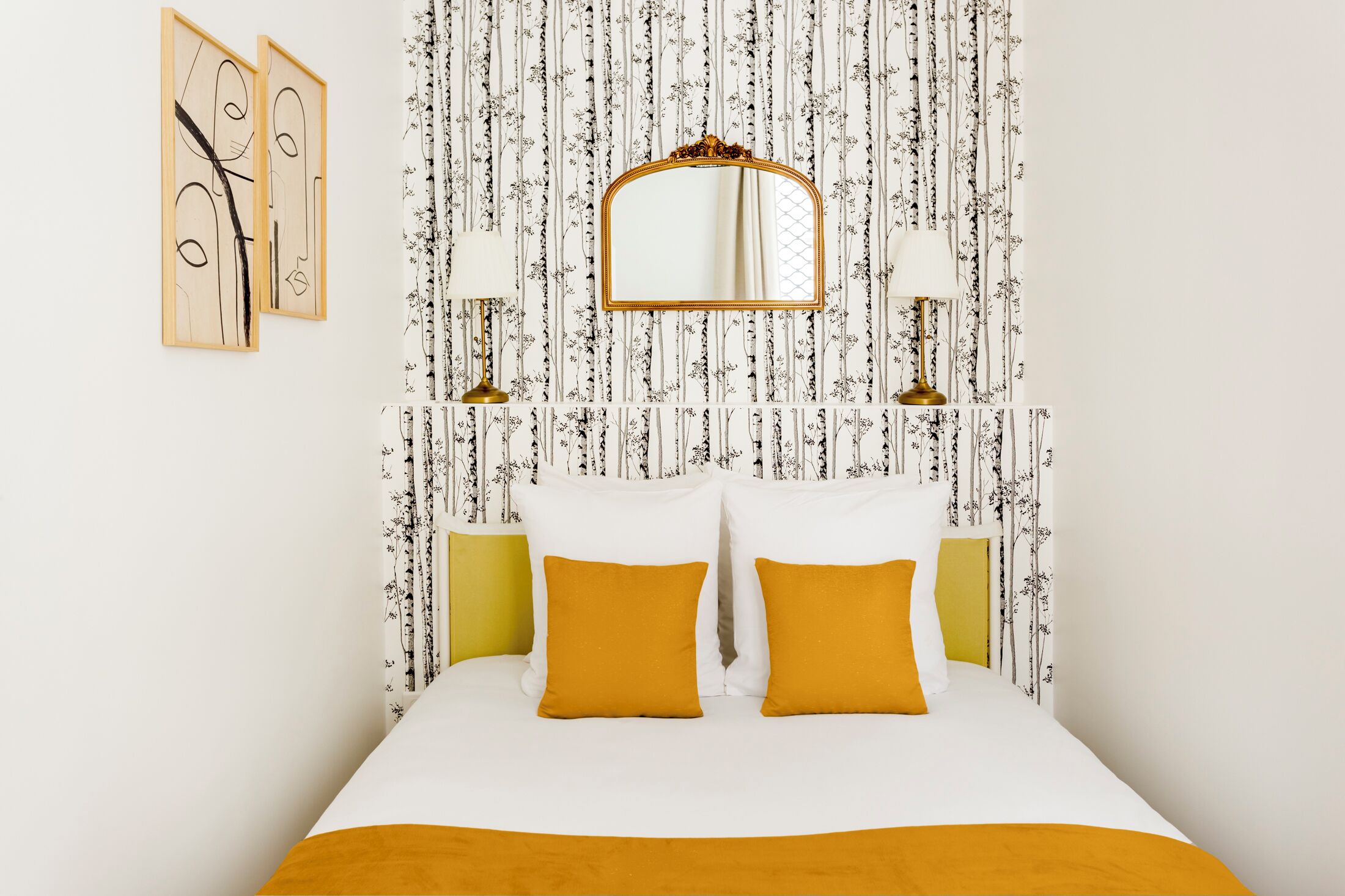 Une chambre à coucher bien rangée, avec un lit, deux lampes de chevet, un miroir et des œuvres d’art accrochées au mur.
