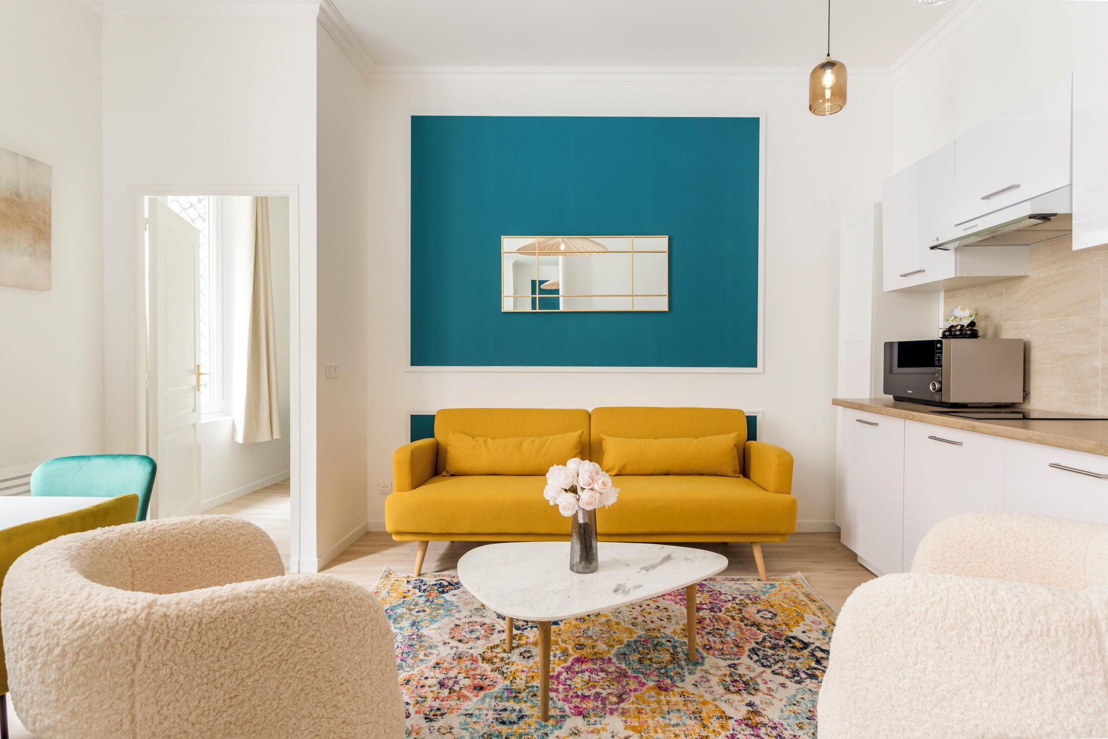 A modern living room with a yellow sofa, a white coffee table with a vase of flowers, a colorful rug, and a large blue wall with a mirror.