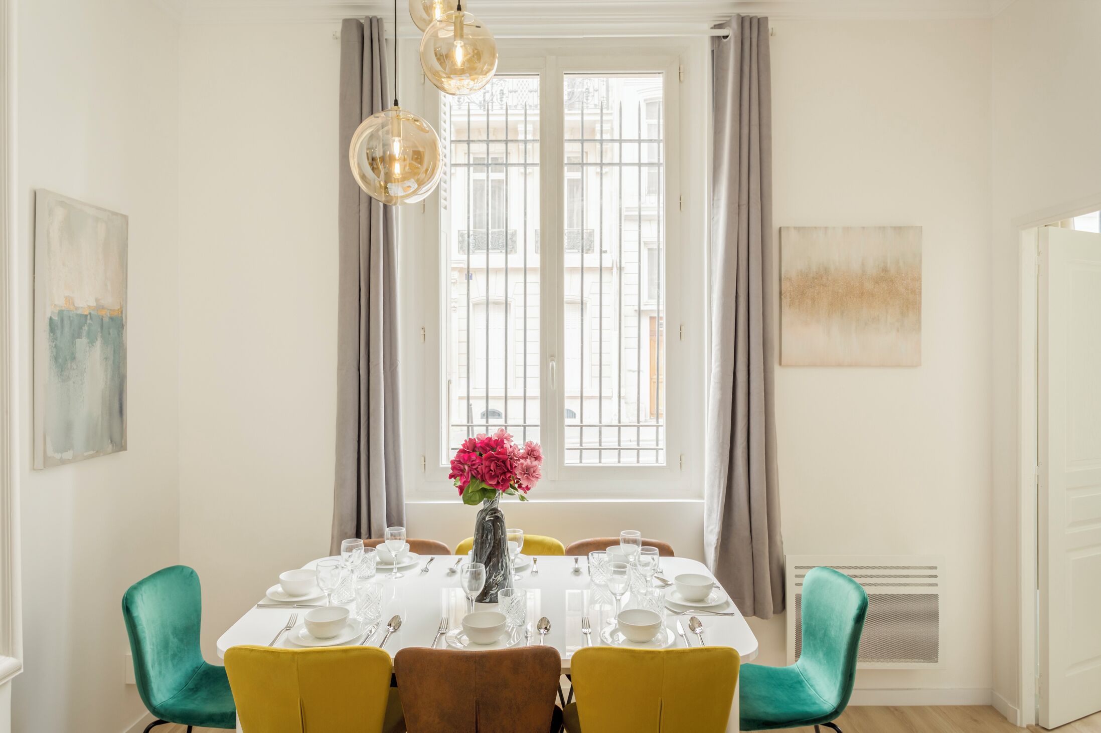 A dining room with a white table set for six, yellow chairs, a vase with pink flowers, and a window with curtains.