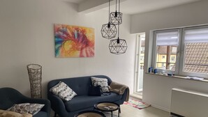 A modern living room with a blue sofa, two armchairs, a coffee table, a colorful abstract painting, and a large window with a view of buildings.