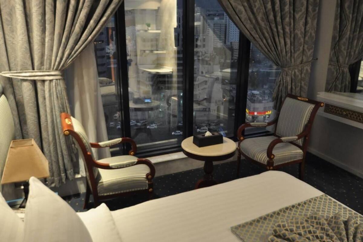 Een hotelkamer met een groot raam, een bed, twee stoelen, een klein tafeltje en een lamp.
