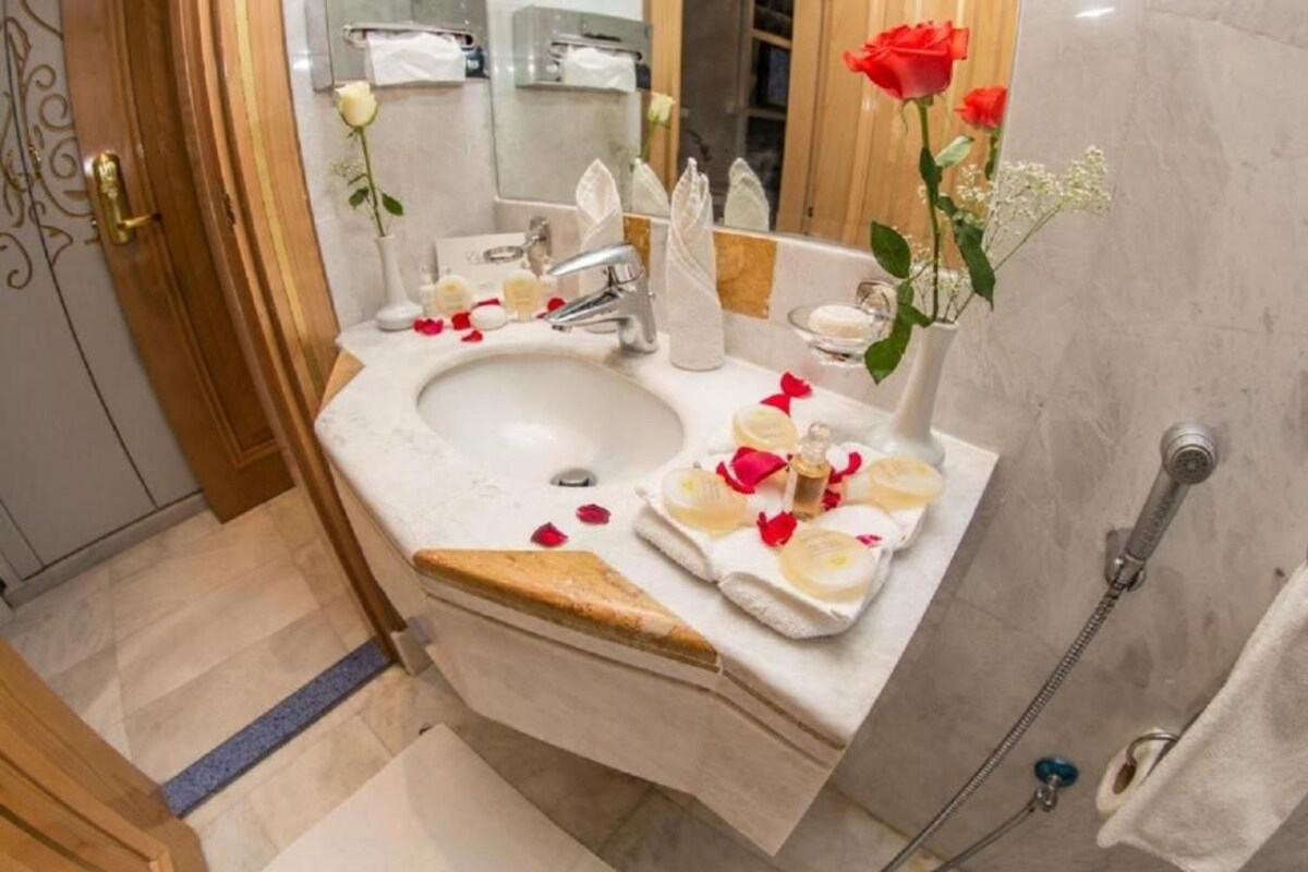 Une salle de bain avec un lavabo, un miroir et une douche. On y trouve des pétales de rose et un vase rempli de roses sur le comptoir.