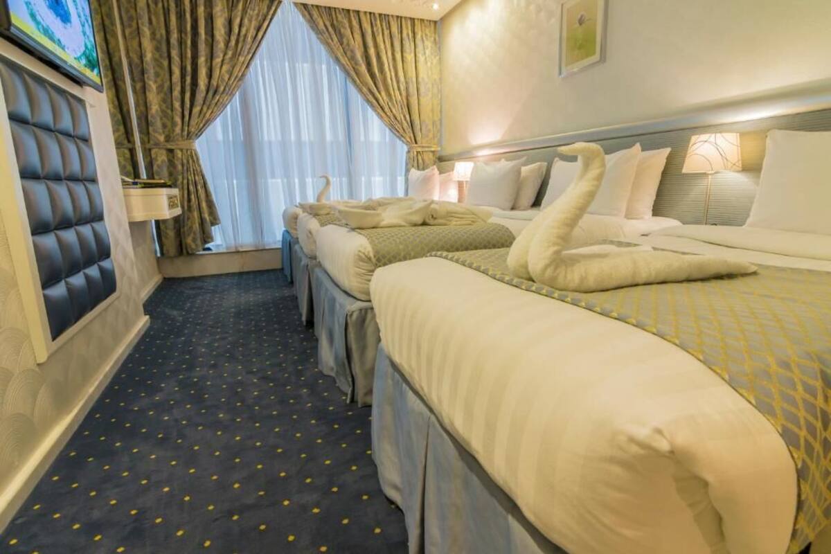 Een hotelkamer met een groot bed, twee kleinere bedden, een bekleed hoofdbord, een raam met gordijnen, en een vloerkleed met een patroon.