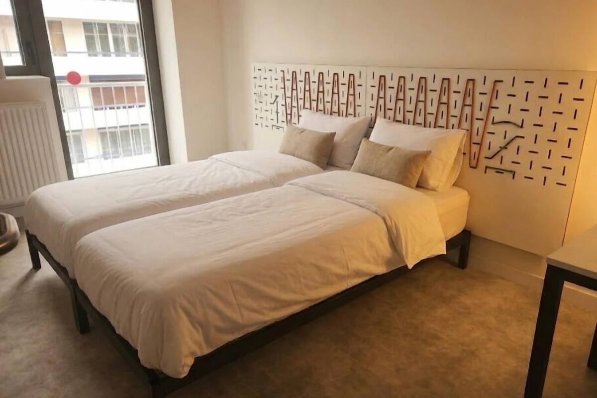 Ein Doppelbett mit einem Kopfende mit geometrischem Muster, ein Fenster mit Blick auf ein Gebäude und ein Heizkörper auf der linken Seite.