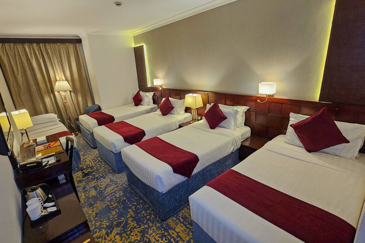 Une chambre d’hôtel avec quatre lits simples, chacun agrémenté d’une couverture rouge et de coussins, un bureau avec une lampe, une télévision et un minibar.
