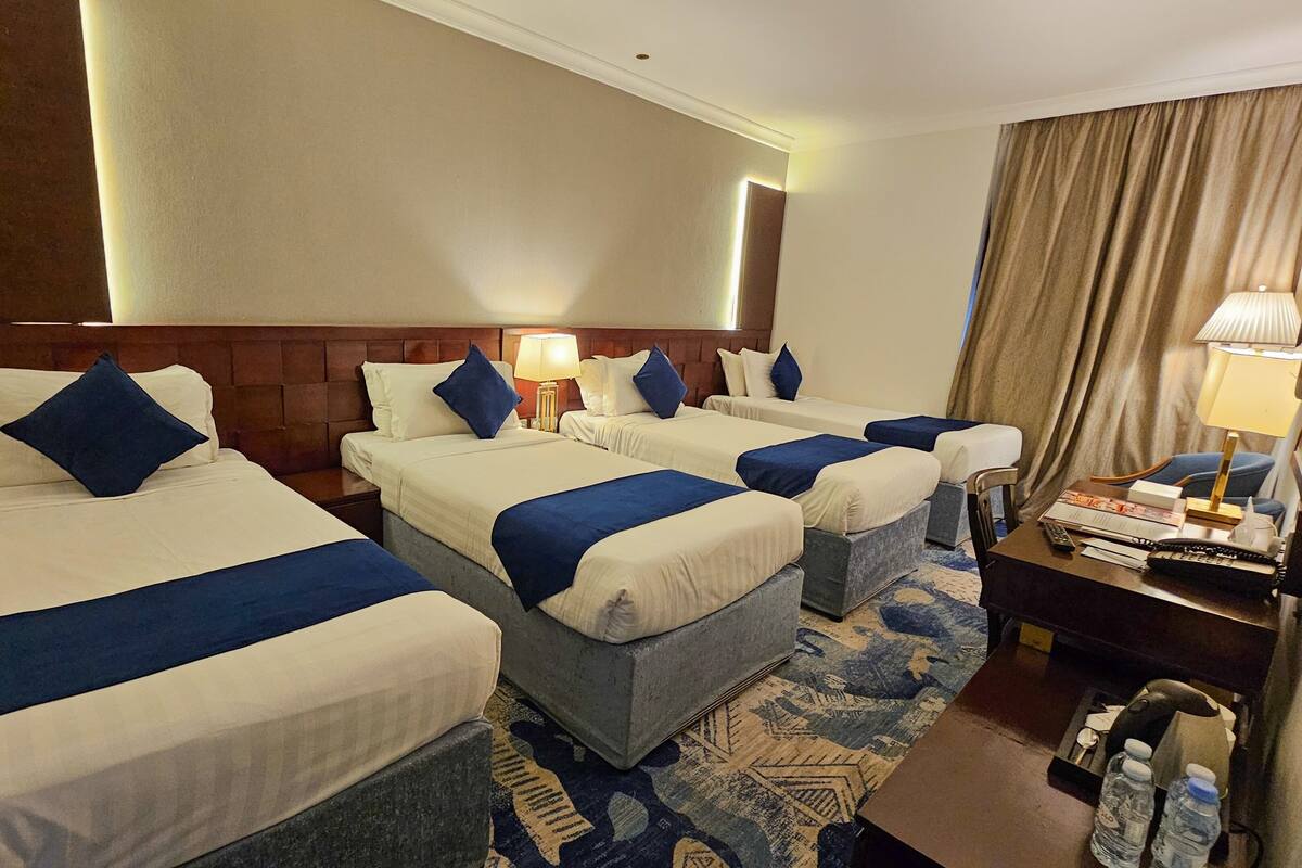 Une chambre d’hôtel avec trois lits, chacun agrémenté de draps blancs et de touches de bleu, un petit bureau avec une lampe et une table de chevet avec un téléphone et des bouteilles d’eau.