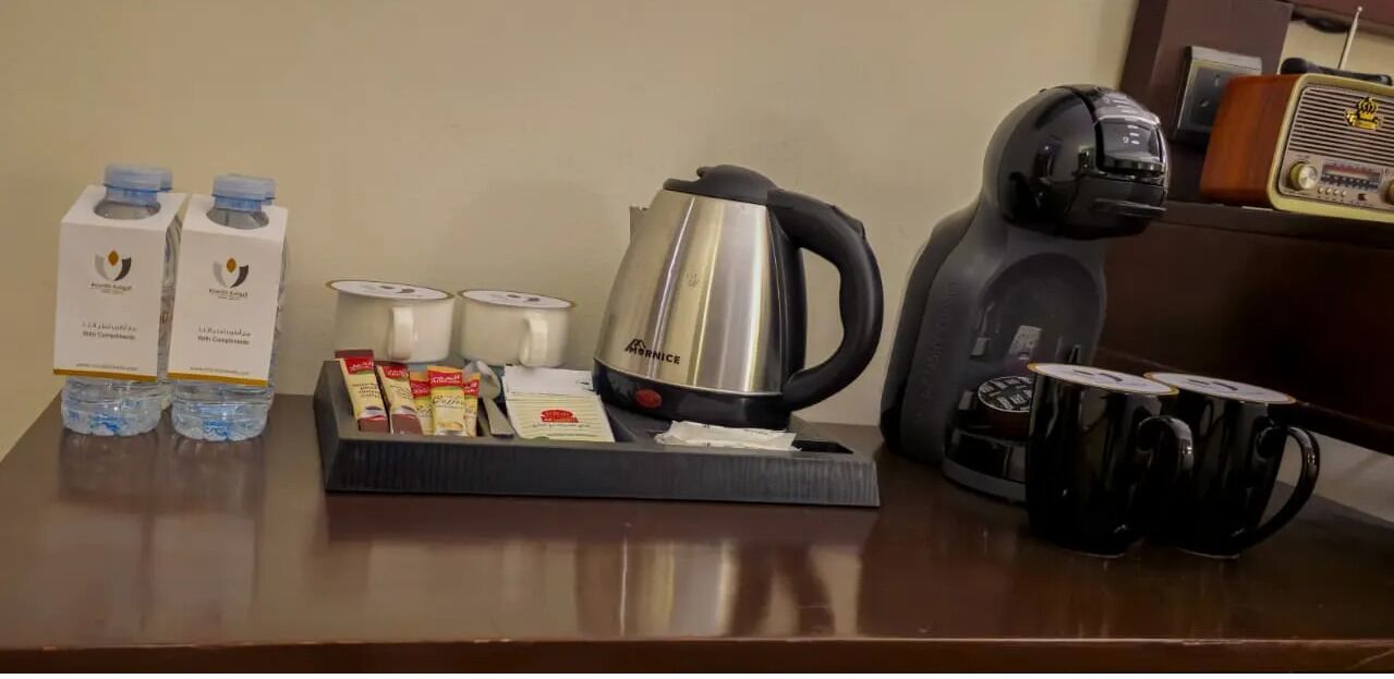 A hotel room's breakfast setup with a coffee maker, tea kettle, water bottles, and a tray of food items.