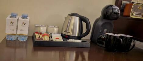 A hotel room's breakfast setup with a coffee maker, tea kettle, water bottles, and a tray of food items.