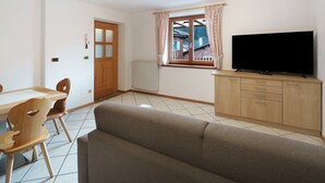 A living room with a sofa, wooden chairs, a television on a cabinet, a window with curtains, and a door.